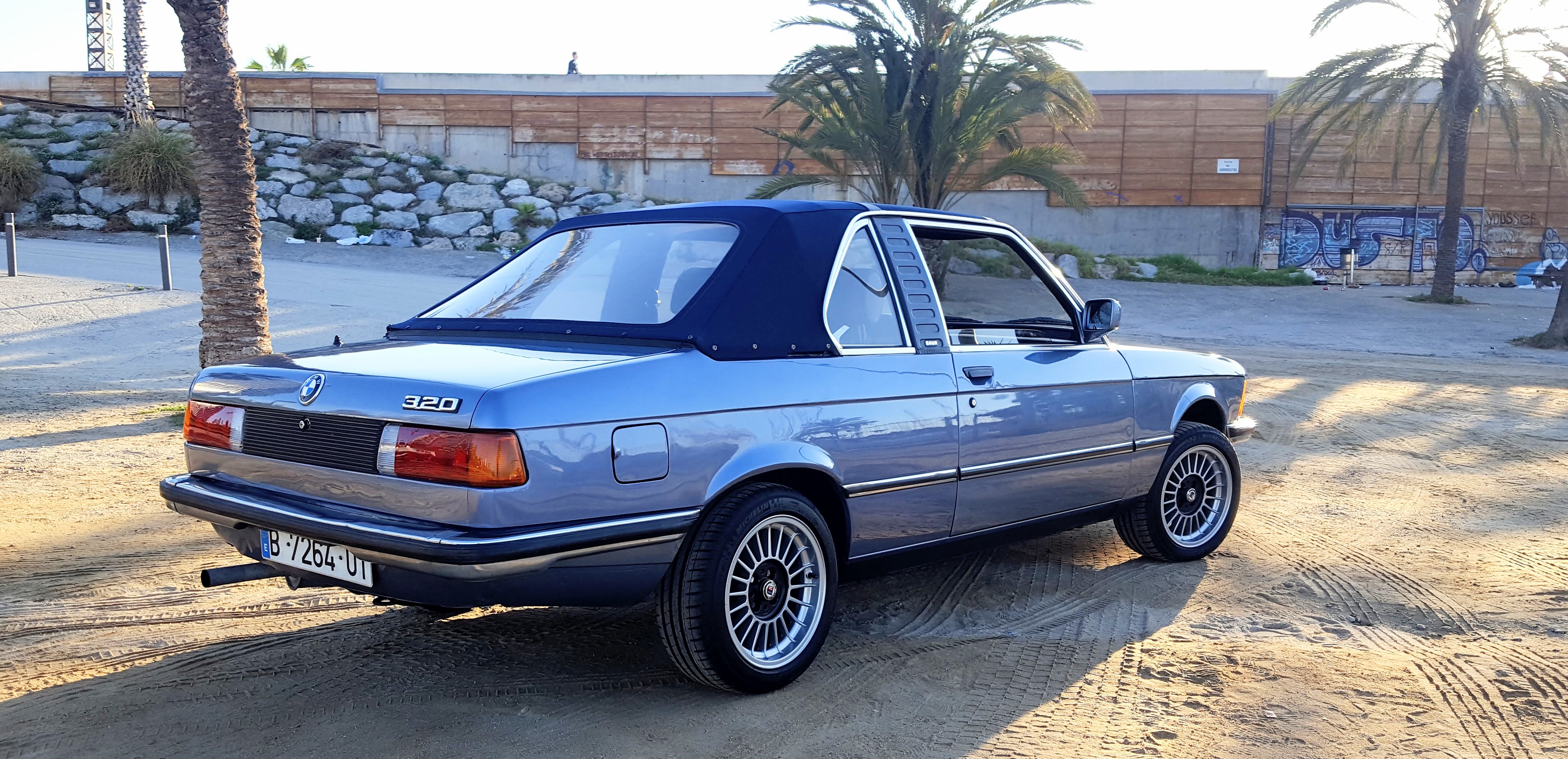 Vista trasera tres cuartos del BMW E21 en una playa de Barcelona con palmeras — escenario para videoclip